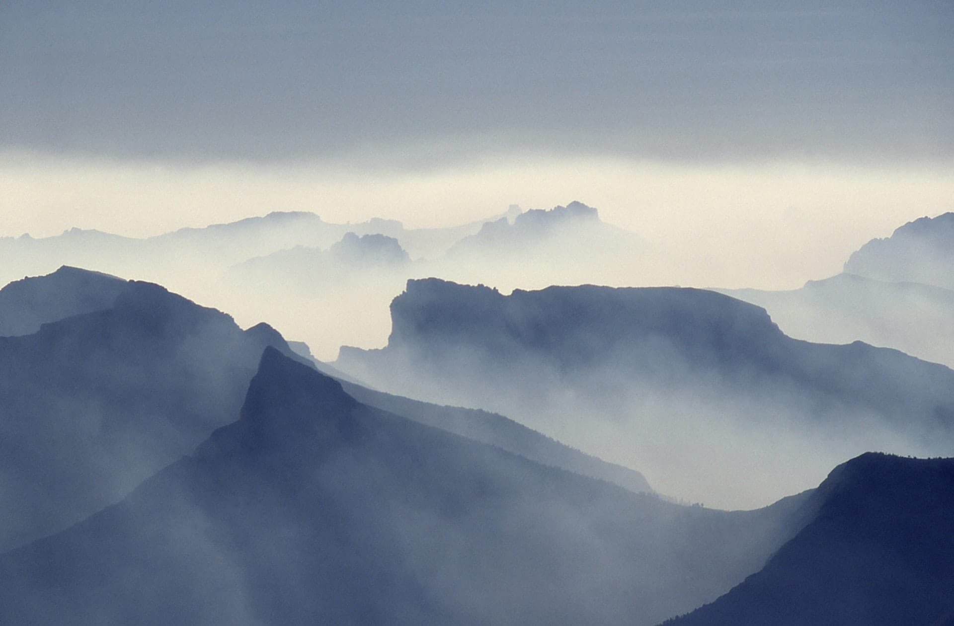 In der Ferne verschwinden mehrere Bergkämme, umhüllt von Nebel und sanftem blaugrauen Licht. Die Szene wirkt ruhig und ätherisch, mit sanften Abstufungen zwischen den einzelnen Bergkämmen, die eine verträumte, stimmungsvolle Landschaft in der Morgen- oder Abenddämmerung schaffen.
